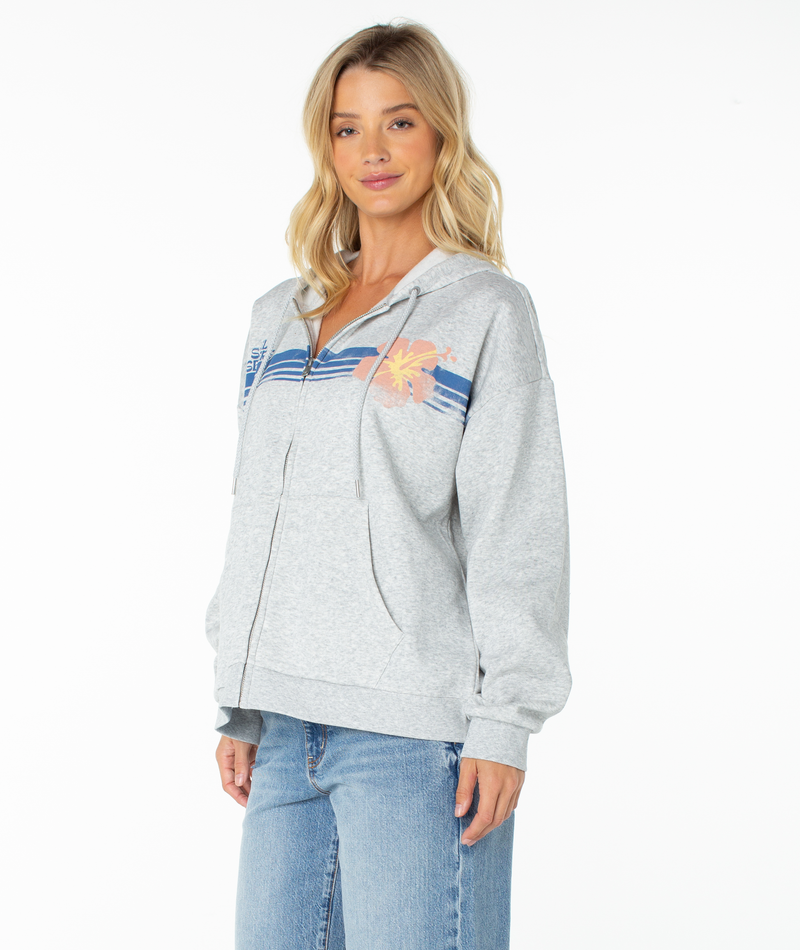 A woman stands against a plain white background wearing the Roxy Sundown Zip Hoodie, featuring a buttery soft light gray fabric with a blue stripe and orange flower design, paired with light blue jeans.