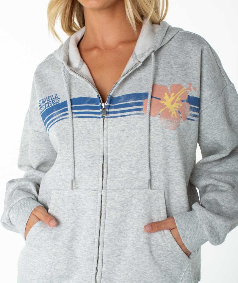 A woman wears the Roxy Sundown Zip Hoodie—a light gray zip-up with blue stripes, a small chest logo, and a peach hibiscus flower design. Stylish and buttery soft, it’s perfect beachwear with comfy pockets for her hands.