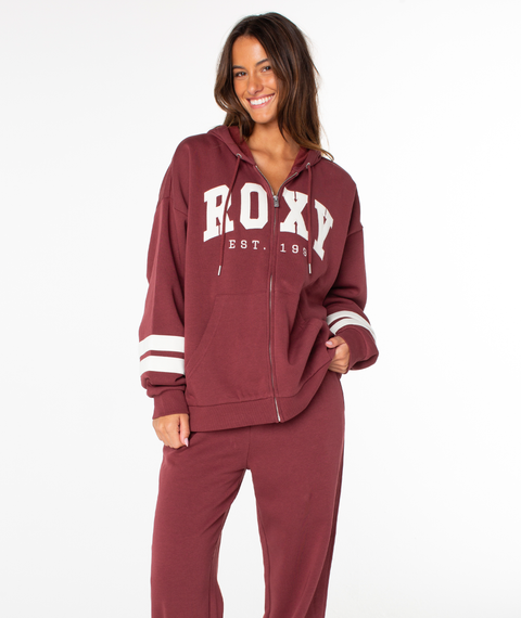 Wearing the Roxy Sunset Zip Top in the rich Port End Game shade, featuring a bold ROXY graphic and “ROXY EST. 1990” printed on the front, this woman pairs it with matching pants and smiles against a plain white background, radiating vintage vibes.