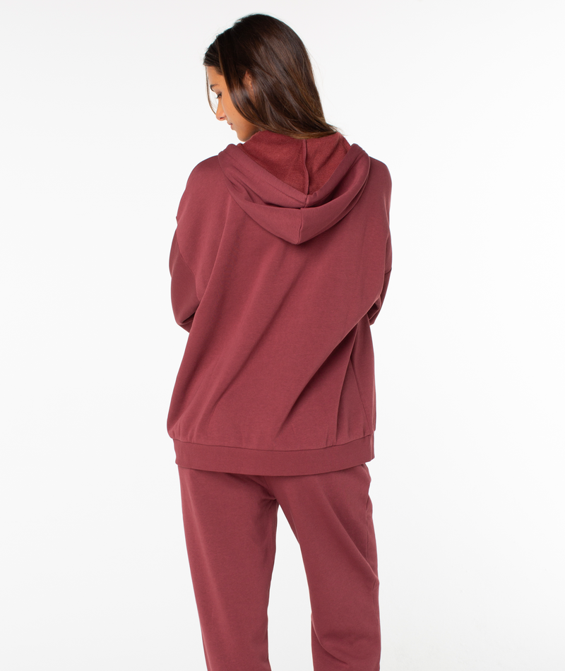 A person with long brown hair stands facing away, wearing the Roxy Sunset Zip Top in maroon with vintage vibes, against a plain white background.