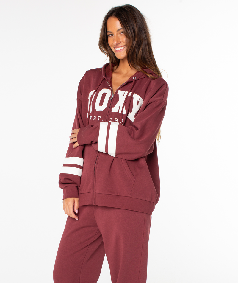 A woman in a maroon Roxy Sunset Zip Top and matching pants stands smiling, with one arm crossed over her body. The zip-up hoodie features bold white stripes on the sleeves and a large ROXY graphic across the chest, channeling vintage vibes reminiscent of a retro varsity jacket. The background is plain white.