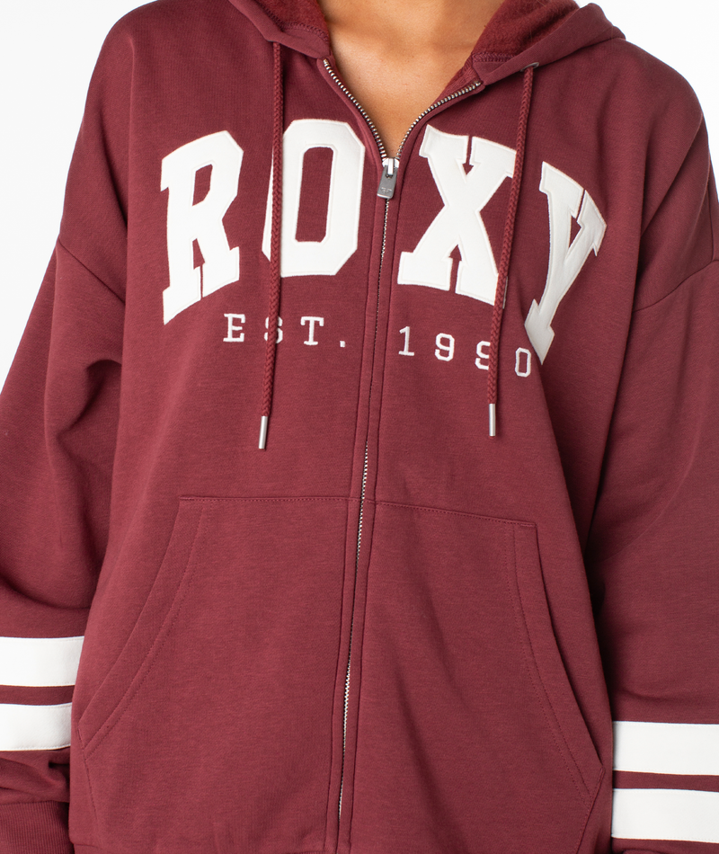 A person is wearing the Roxy Sunset Zip Top—a maroon zip-up hoodie with a bold white ROXY graphic and EST. 1990 printed on the front. Featuring white stripes on the sleeves and drawstrings on the hood, this hoodie channels vintage vibes inspired by classic retro varsity jackets.