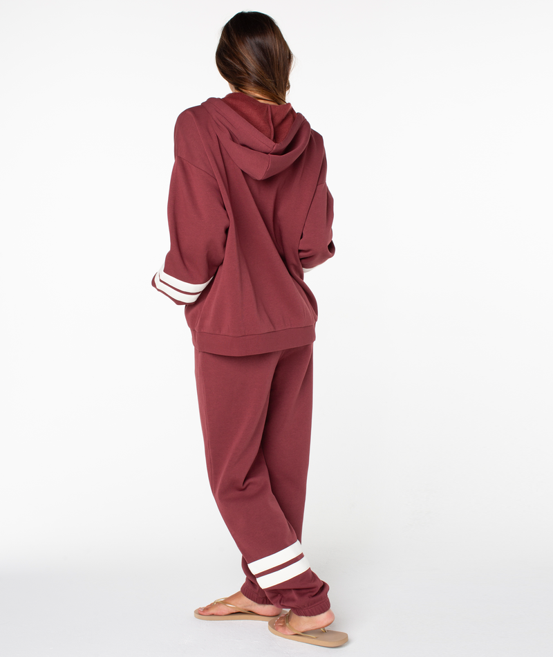 A person stands barefoot with their back to the camera, wearing the loose-fitting Roxy Sunset Zip Top in maroon, featuring a subtle ROXY graphic, paired with matching pants detailed with white stripes near the cuffs, all set against a plain white background for an effortless vintage vibe.