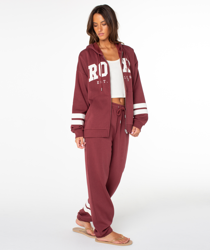 A woman wearing the Roxy Sunset Zip Top, a maroon hooded sweatshirt with a bold ROXY graphic, and matching sweatpants stands on a white background, looking down with one hand in her pocket and tan flip-flops on her feet.