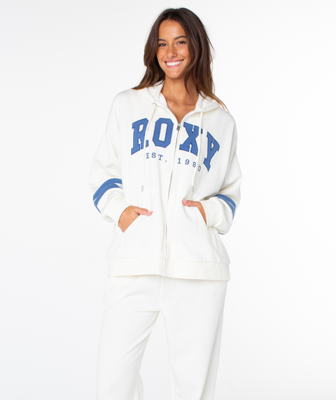 A woman with long brown hair smiles while wearing the Roxy Sunset Zip Top in Cloud Dancer End Game, paired with matching white pants, standing against a plain white background.