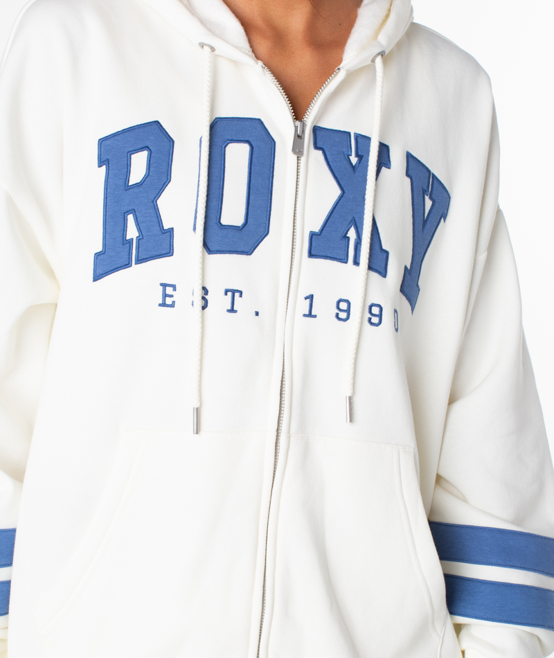 A person wears the Roxy Sunset Zip Top, a white vintage-style zip-up hoodie with blue ROXY graphic letters and "EST. 1990" on the front, drawstrings, and blue double sleeve stripes for a retro varsity look.