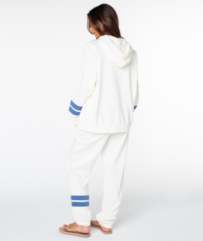 A person faces away, wearing the Roxy Sunset Zip Top—a retro-inspired off-white hoodie with blue stripes—paired with matching sweatpants and beige flip-flops against a plain white background.