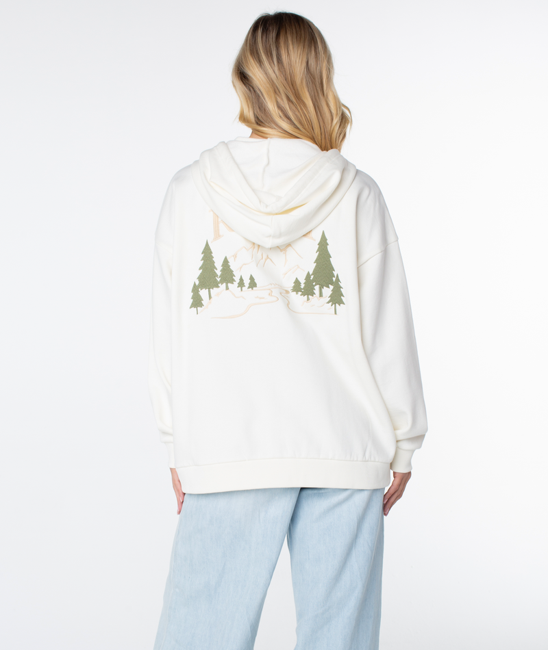 A person with long, wavy blonde hair wears the Roxy Line Up Zip hoodie with a mountain and pine tree graphic on the back, paired with light blue jeans, standing against a plain white background.