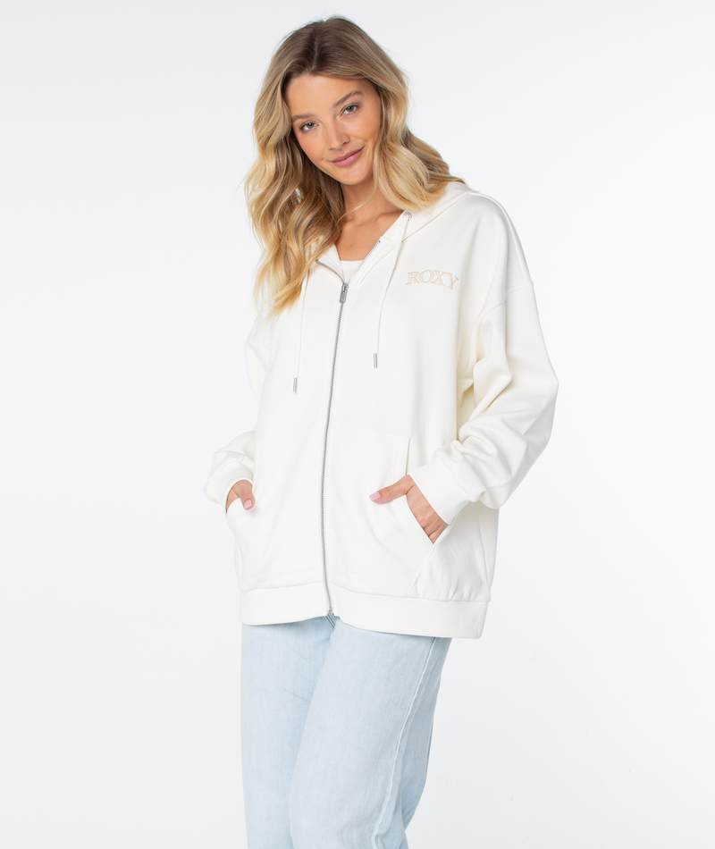 A woman with long blonde hair models the Roxy Line Up Zip white cotton blend hoodie and light blue jeans, standing with her hands in her pockets against a plain white background.