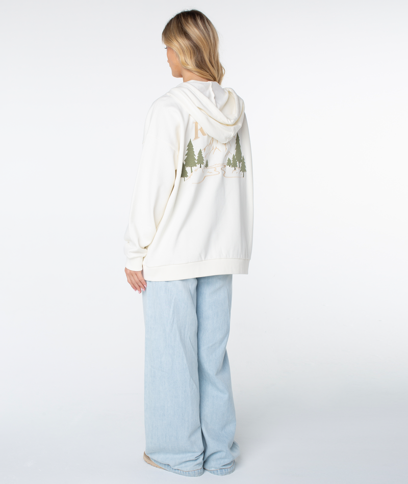 A person with long blonde hair, wearing the Roxy Line Up Zip cotton blend hoodie with a scenic back print and light blue wide-leg jeans, stands facing away in a bright studio.