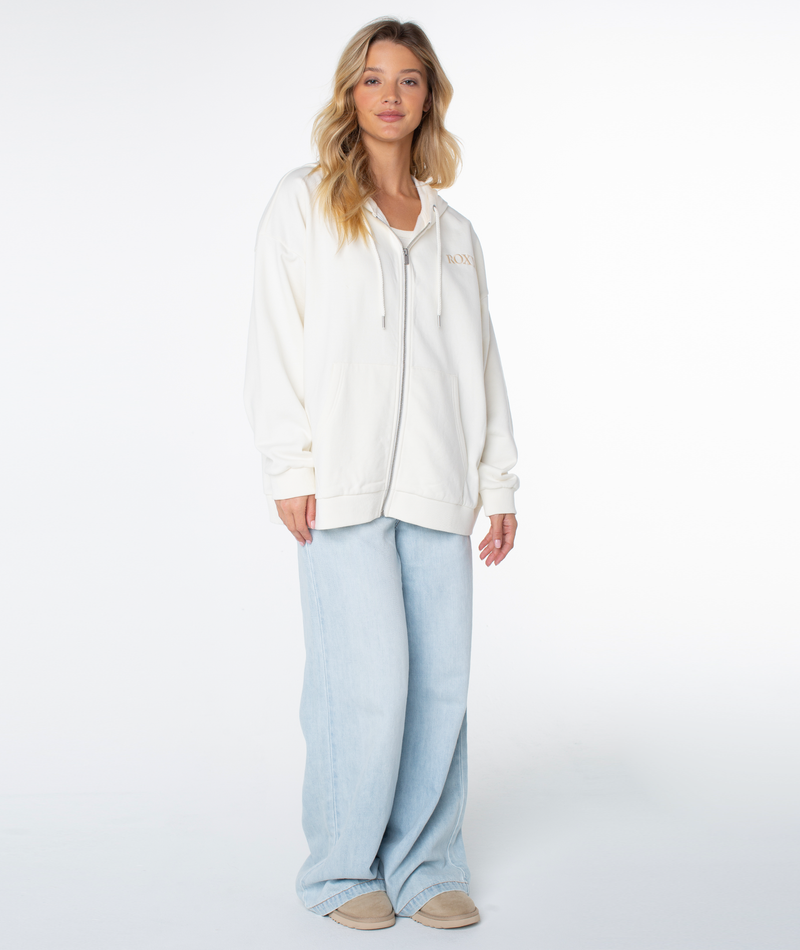 A person stands against a white background in a relaxed style, wearing the Roxy Line Up Zip hoodie in Coconut Milk Mt Embroidery, paired with light blue wide-leg jeans and beige shoes. Their long wavy blonde hair frames their face.