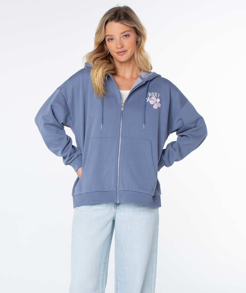 A blonde woman wears the Roxy Line Up Zip, a blue cotton blend zip hoodie with floral design, paired with light-wash jeans. She stands hands on hips against a white background, showing her casual style.