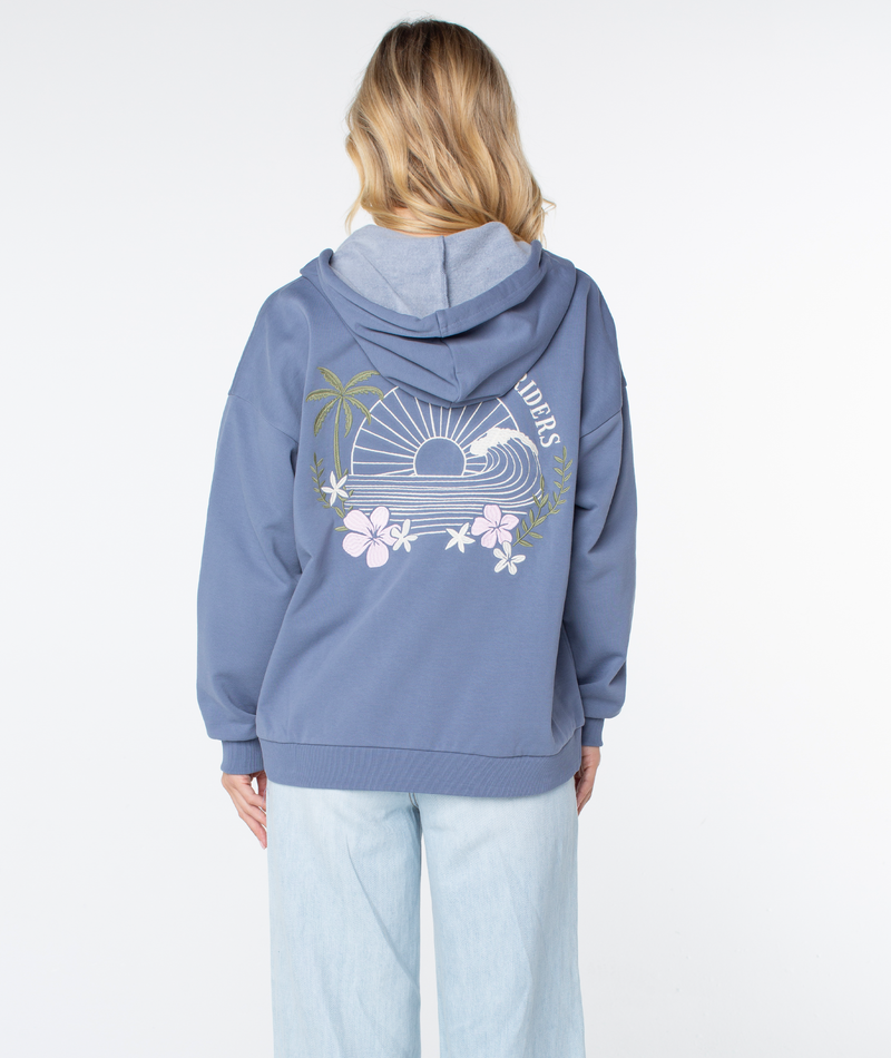 A person with long blonde hair wears the Roxy Line Up Zip—a blue cotton blend zip up hoodie with a sunset, palm trees, and flowers on the back—paired with light blue jeans against a plain white background.