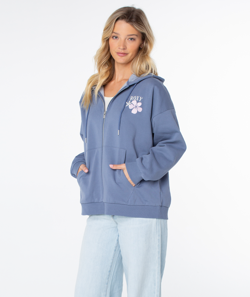 A person wears the Roxy Line Up Zip—a blue cotton blend zip-up hoodie with a small chest graphic—paired with light-wash jeans, standing hands-in-pockets against a plain white background.