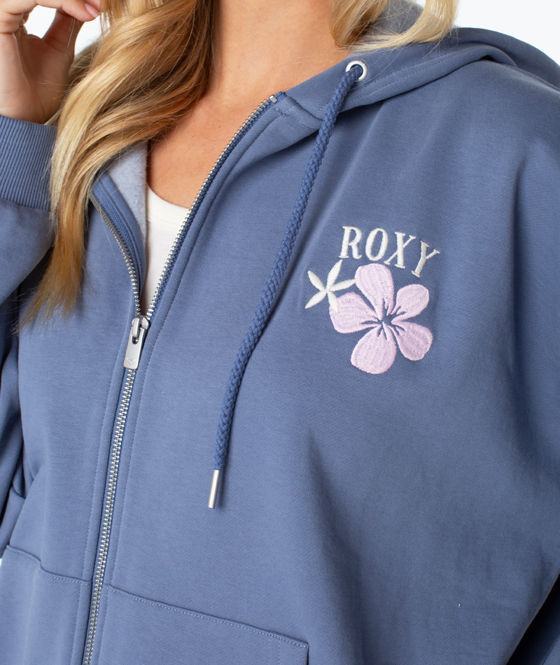 A person wears the Roxy Line Up Zip in blue cotton blend with a white drawstring, embroidered ROXY text, and a pink flower with white star on the left chest; only part of their face is visible.