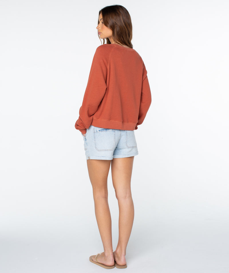 A woman stands facing away, wearing a loose rust-colored Roxy Golden Hour Top, light denim shorts, and tan sandals against a plain white background, embodying an effortless sense of coastal comfort.
