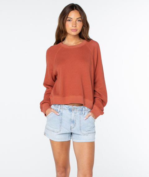 A person stands against a plain white background wearing the Chutney Solid Golden Hour Top by Roxy, crafted from lightweight cotton and paired with light denim shorts, hands in their pockets, embodying effortless coastal comfort.