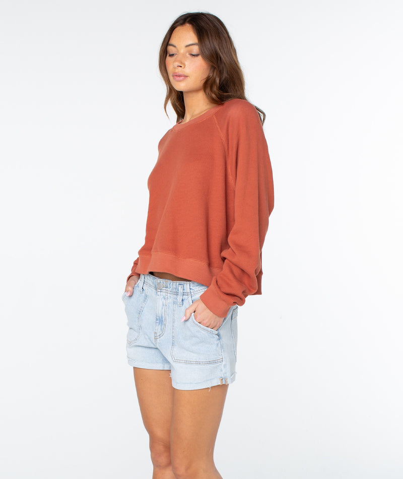 A woman with long brown hair stands with her hands in her pockets against a plain white background, embodying coastal comfort in the laidback, lightweight cotton style of the rust-colored Roxy Golden Hour Top paired with light denim shorts.