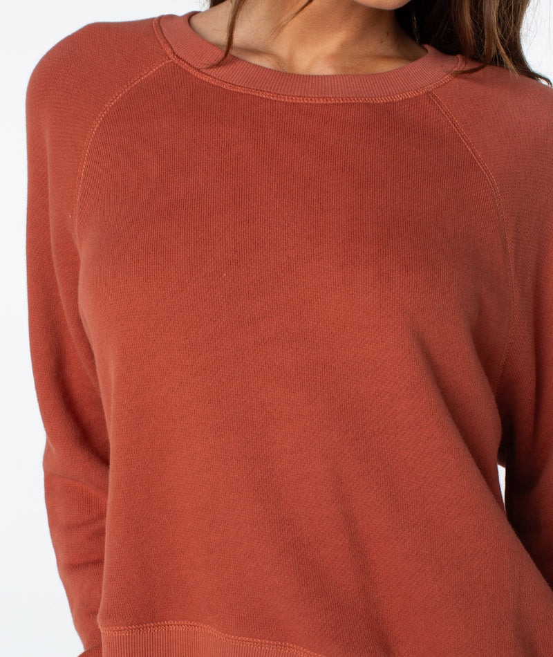A person wears the Roxy Golden Hour Top—a long-sleeved, rust-colored sweatshirt made from lightweight cotton. With only their upper torso visible against a plain, light background and their face partially out of frame, the image conveys an effortless sense of coastal comfort.