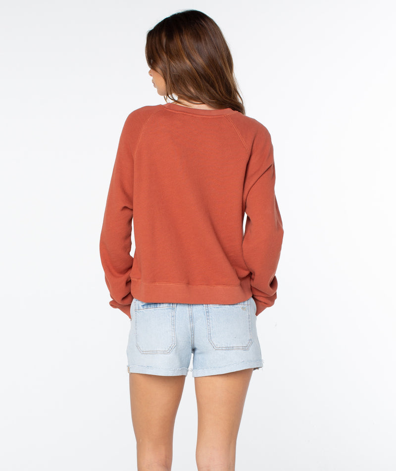 A person with long brown hair faces away, wearing the rust-colored Roxy Golden Hour Top crafted from lightweight cotton and light denim shorts, standing against a plain white background and evoking an effortless sense of coastal comfort.