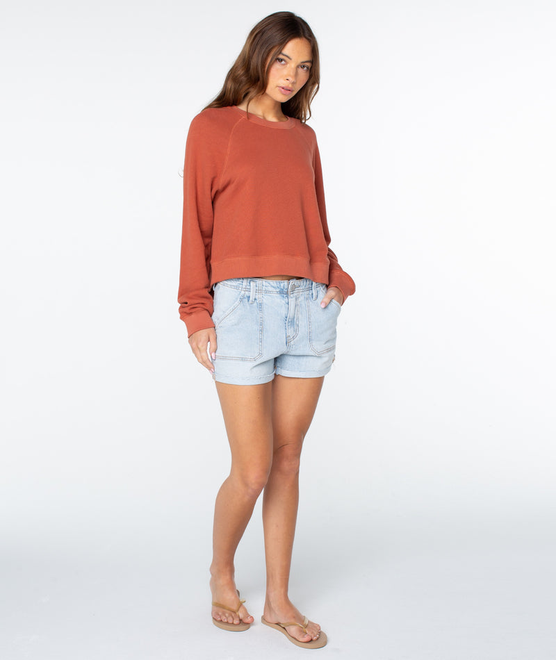 A woman stands against a plain background, wearing the rust-colored Golden Hour Top by Roxy—a lightweight cotton long-sleeve style—paired with light denim shorts and beige flip-flops. With one hand in her pocket and a relaxed expression, she perfectly captures effortless coastal comfort.
