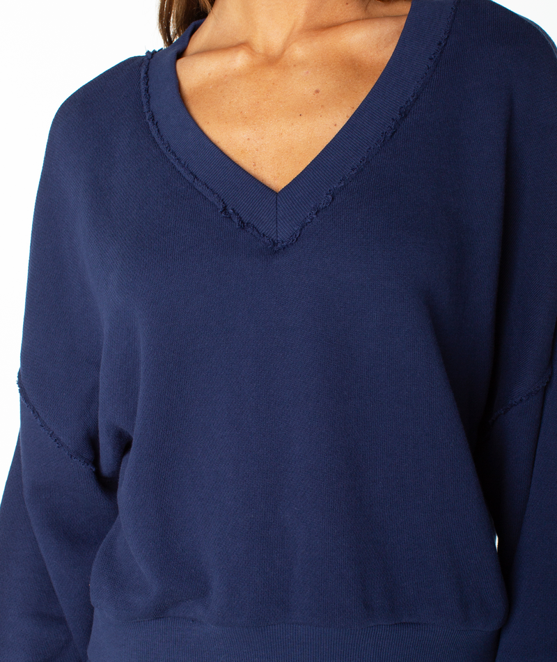 A person wearing the Roxy Dream Thing Top—a navy blue, long-sleeve V-neck sweater featuring raw-edge details and a relaxed loungewear vibe. The photo is cropped from shoulders to waist, with no visible background.