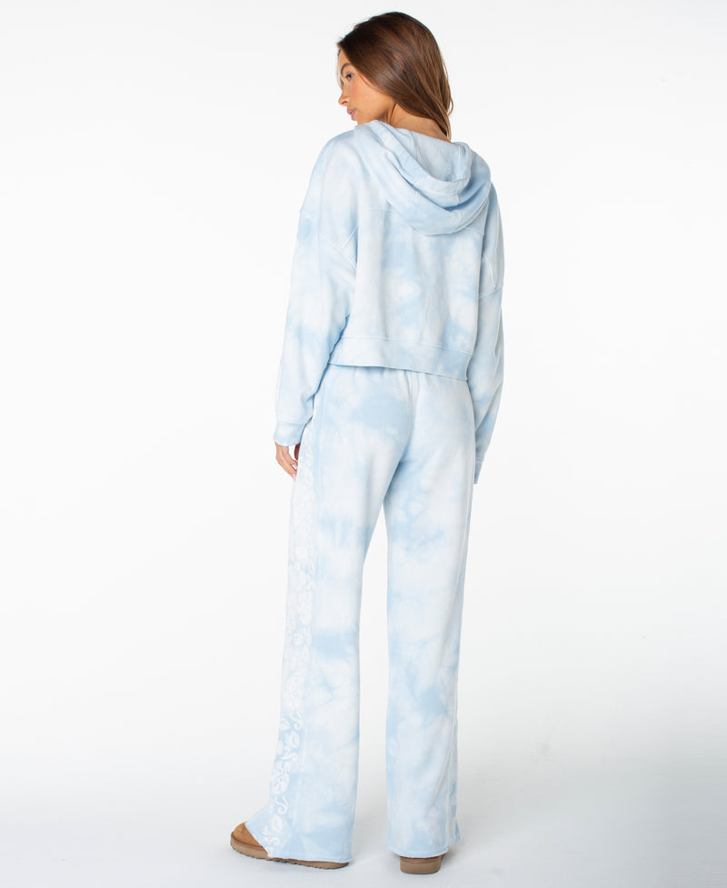A person stands facing away, wearing the Roxy Fringing Reefs Top in a light blue and white tie dye pattern with matching loungewear pants. The outfit has a relaxed fit, and the person is wearing tan shoes. The background is plain white.