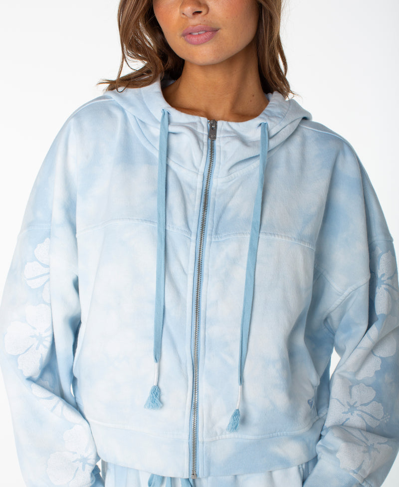 A woman is wearing the Roxy Fringing Reefs Top, a light blue tie-dye zip-up hoodie with a hood and drawstrings. This cozy piece of loungewear features delicate floral patterns on the sleeves. The image is cropped at the chin and above the knees.