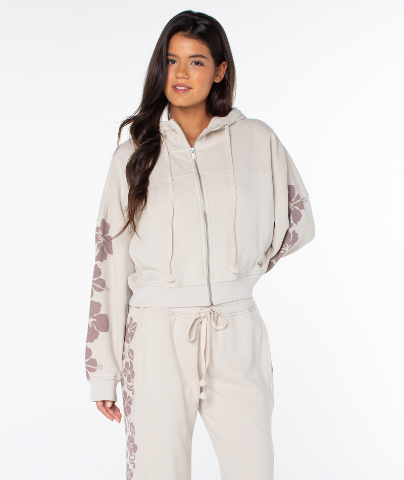 A woman wears the Roxy Fringing Reefs Top—a light beige zip-up hoodie with floral sleeve prints—paired with matching beige sweatpants, standing against a white background.