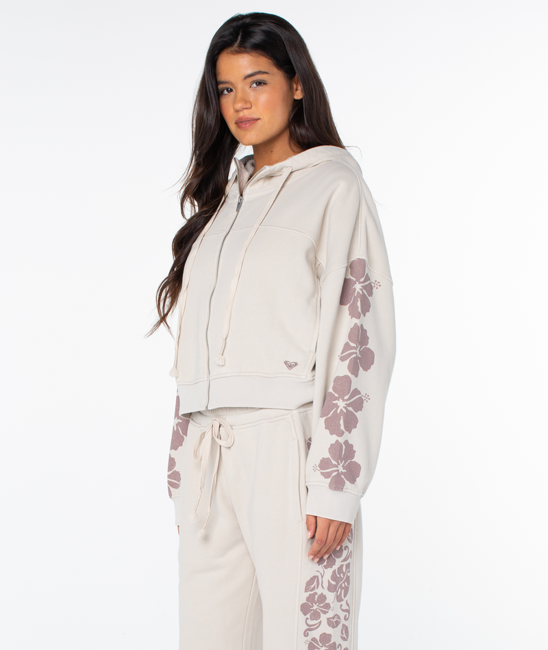 A woman wears the Roxy Fringing Reefs Top, a light beige zip-up hoodie with floral prints on the sleeves, standing against a plain white background.