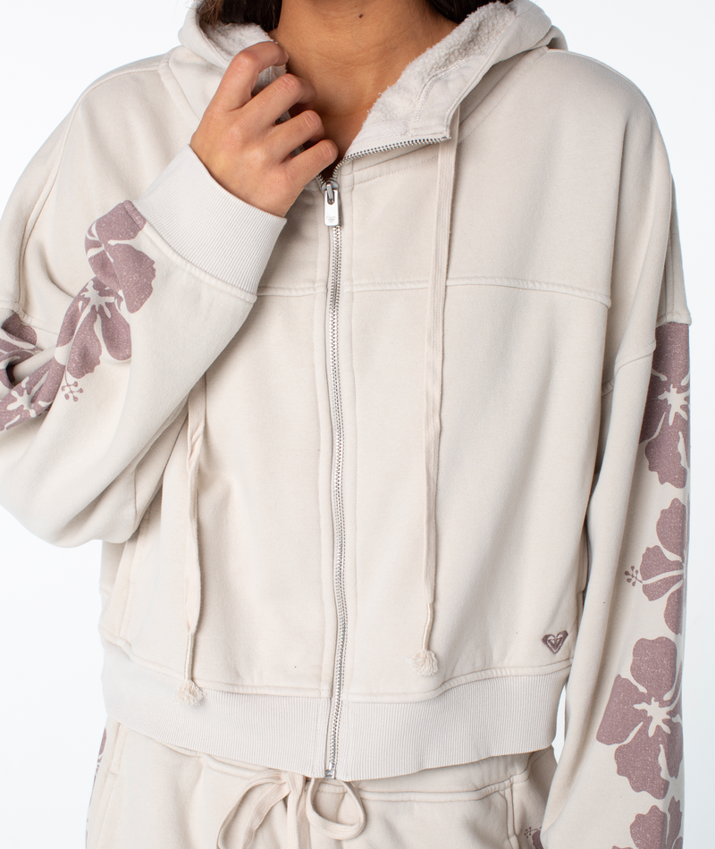 A person wears the Roxy Fringing Reefs Top, a light beige zip-up hoodie with floral sleeve prints and drawstrings, paired with matching pants, holding the hoodie near their chin.