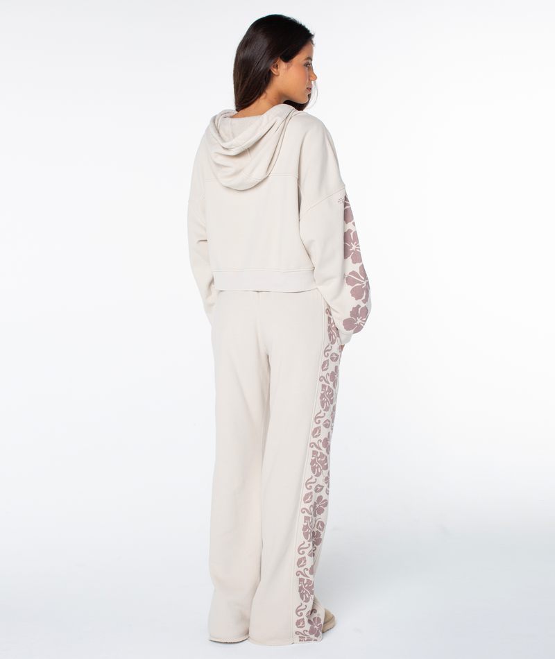 A person stands facing away, wearing the Roxy Fringing Reefs Top—a light beige zip-up hoodie with floral patterns on the sleeves—paired with matching wide-leg pants, against a plain white background.