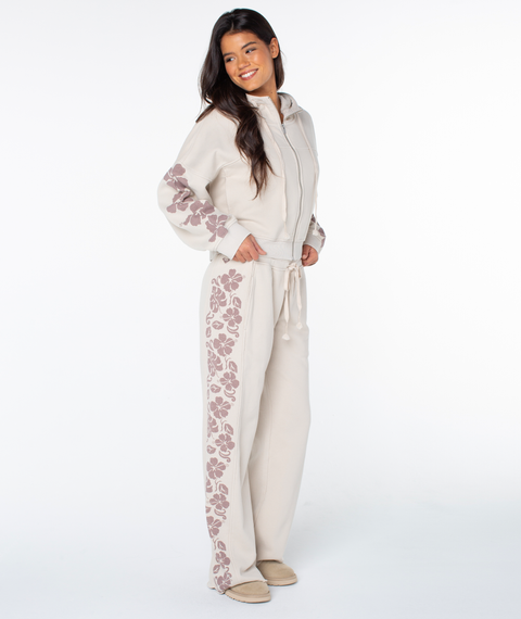 A woman smiles in the Roxy Fringing Reefs Top, color Pumice Stone Fringing Reefs, featuring mauve floral patterns on the sleeves. She wears matching pants and slippers, posing against a plain white background.
