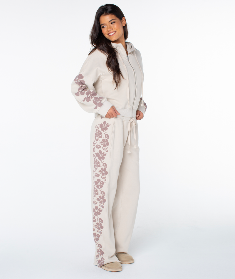 A woman smiles in the Roxy Fringing Reefs Top, color Pumice Stone Fringing Reefs, featuring mauve floral patterns on the sleeves. She wears matching pants and slippers, posing against a plain white background.