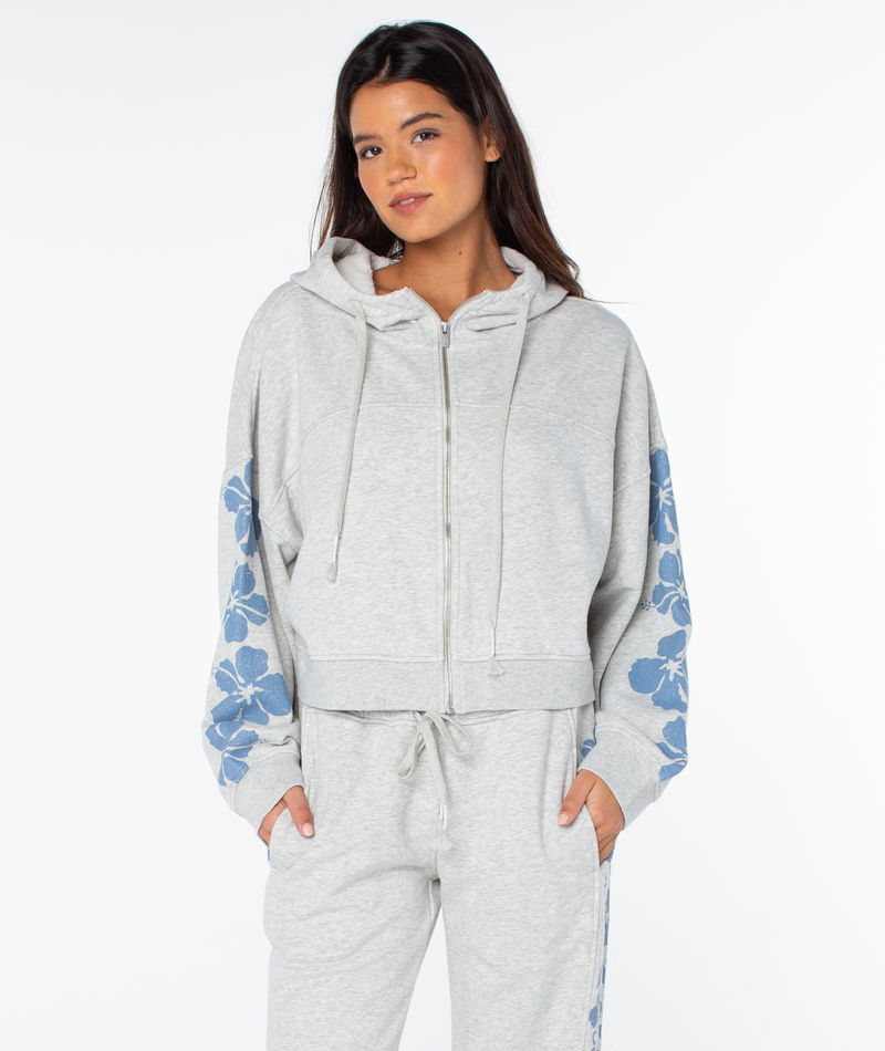 A woman wears the Roxy Fringing Reefs Top, a light gray zip-up hoodie with blue floral sleeve patterns, paired with matching loungewear sweatpants, standing with hands in her pockets against a white background.