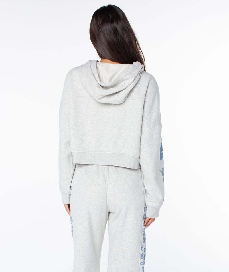 A person with long brown hair wears the Roxy Fringing Reefs Top, a light gray zip-up hoodie with blue floral designs on the sleeves, shown from the back against a white background.