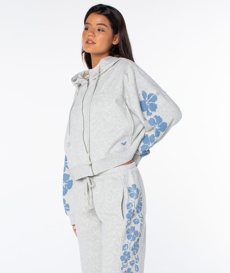A woman wears the Roxy Fringing Reefs Top, a light grey zip-up hoodie with blue floral sleeve patterns, perfect for cozy loungewear, standing against a plain white background.