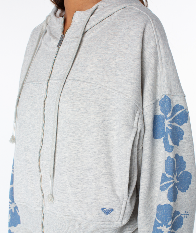 A person wears the Fringing Reefs Top by Roxy, a light gray zip-up hoodie with blue floral sleeve patterns and a small heart logo—ideal loungewear for relaxing days. Only the torso and part of the face are visible.