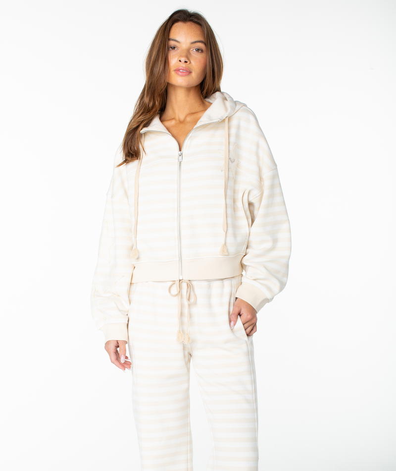 A woman wears the Roxy Fringing Reefs Stripe Top in Parchment Freebird Stripe, paired with matching pants. She stands against a plain white background with one hand in her pocket, looking directly at the camera.