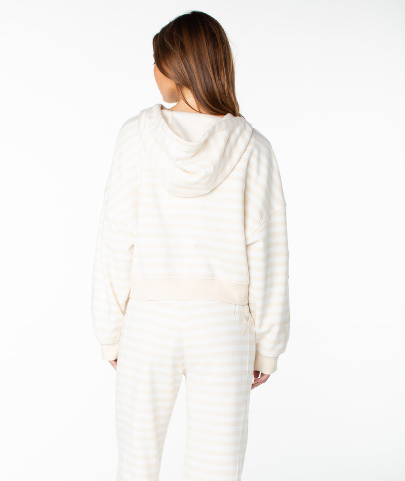 A woman with long brown hair, seen from behind, wears the Roxy Fringing Reefs Stripe Top—a cream and white striped cotton blend hoodie with a boxy fit—paired with matching pants. The background is plain white.