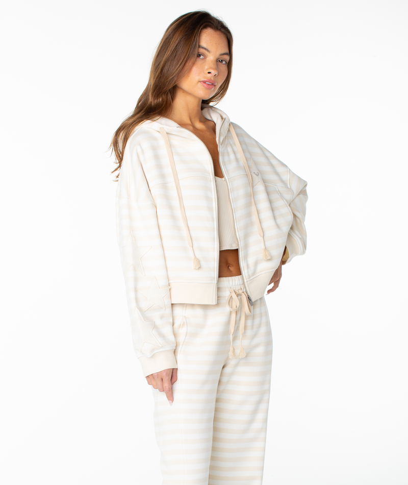 A woman poses against a white background wearing the Roxy Fringing Reefs Stripe Top, a light beige and white striped cotton-blend hoodie with a boxy fit. She has long brown hair and a relaxed, confident expression, pairing the top with matching sweatpants.
