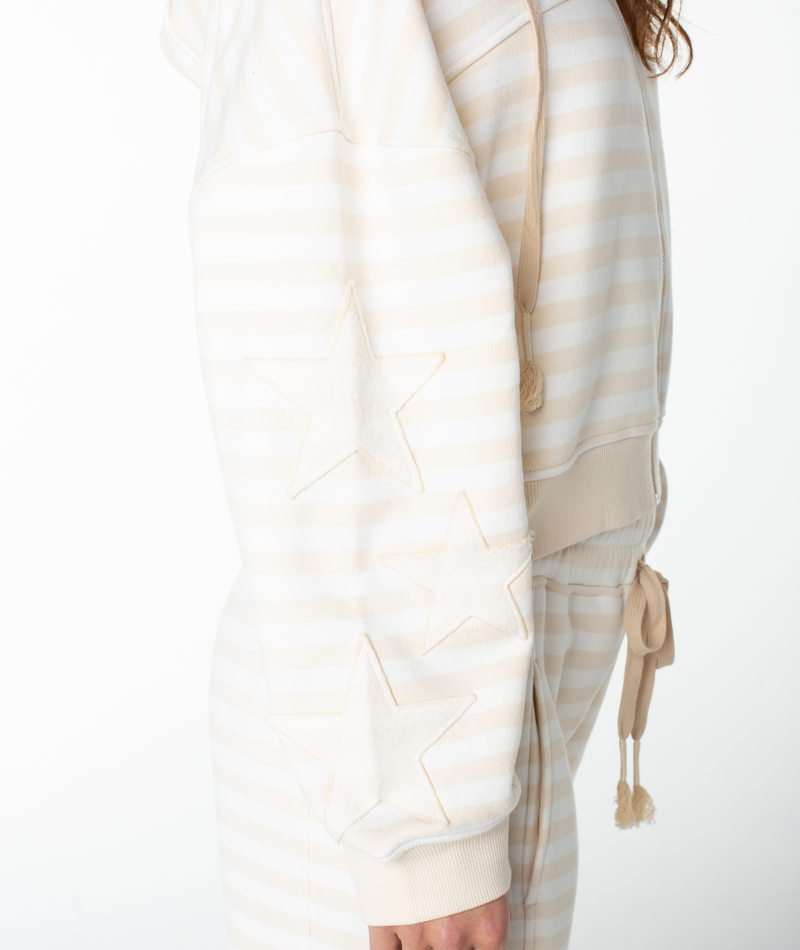 A person is shown from the side against a white background wearing the Roxy Fringing Reefs Stripe Top, a cream and white striped piece featuring two star patches on the sleeve and a boxy fit reminiscent of a cotton blend hoodie.