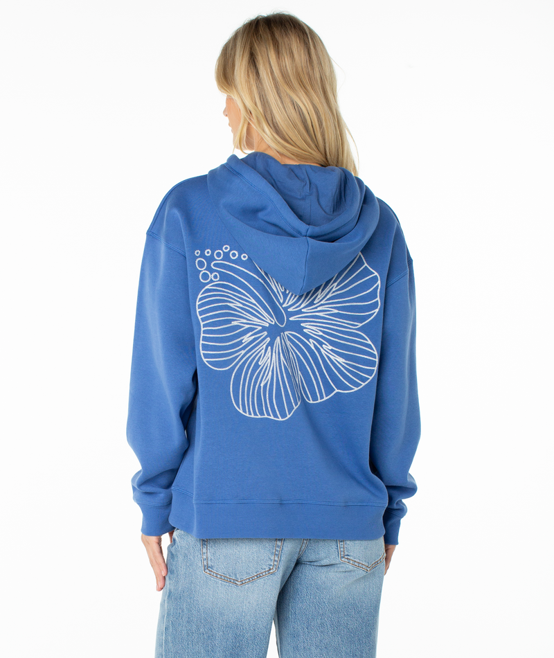 A person with blonde hair wears the Roxy Wailea Hoodie—an ultra-soft blue hoodie featuring a large white abstract flower design on the back—paired with light blue jeans, standing against a white background.