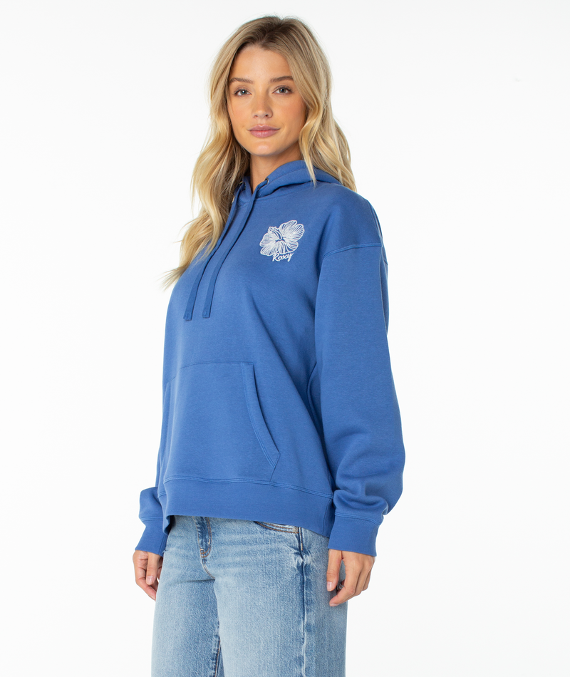 A woman wears the Roxy Wailea Hoodie in blue, adorned with a white floral graphic on the chest, styled with light blue jeans and posed against a plain white backdrop.