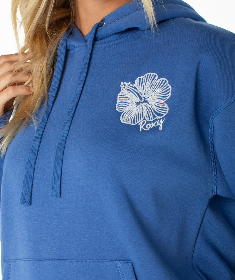 A person is wearing the Roxy Wailea Hoodie, a relaxed blue hoodie with a white hibiscus flower and “Roxy” embroidered on the chest. Only part of their face and hand are visible.