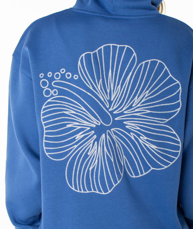 A person wearing a blue Roxy Wailea Hoodie, featuring a large white-outlined hibiscus flower graphic on the back and crafted in an ultra-soft, slouchy hoodie style.
