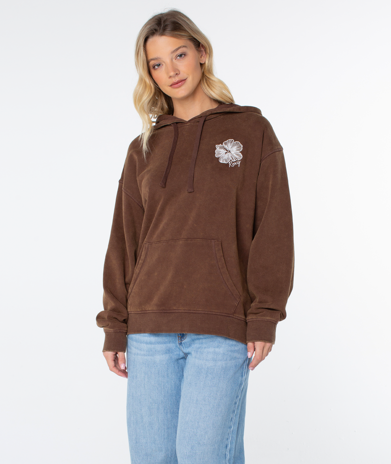 A woman in a Roxy Wailea Hoodie with a flower chest graphic and light blue jeans stands against a white background, looking at the camera.