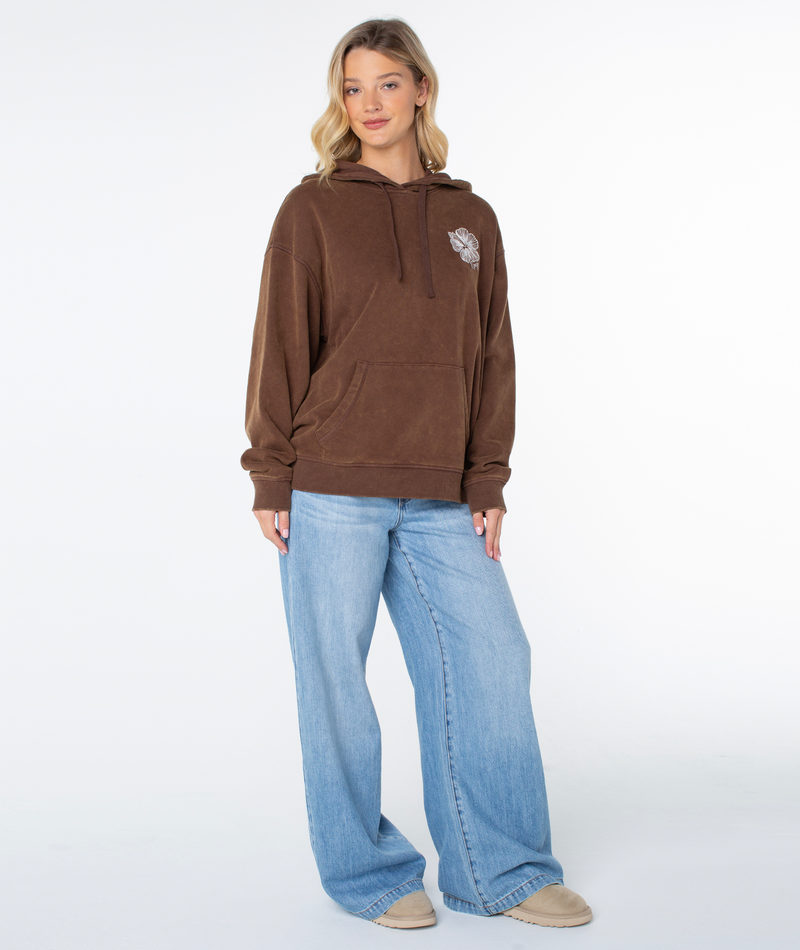 A woman stands facing forward in the Roxy Wailea Hoodie in Chestnut Stitched Hibiscus, featuring a small chest graphic, paired with light blue wide-leg jeans and beige shoes against a plain white background.