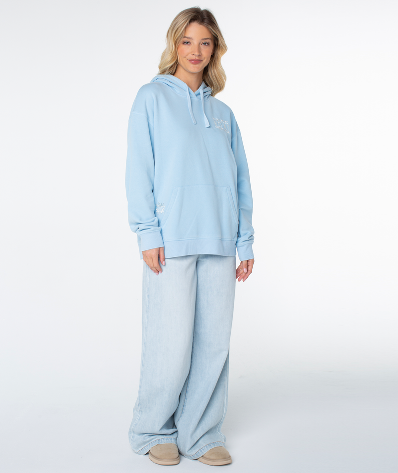 A person smiles, wearing a Roxy Wailea Hoodie in Cerulean Palm Poster with wide-leg light wash jeans and beige shoes against a plain white background.