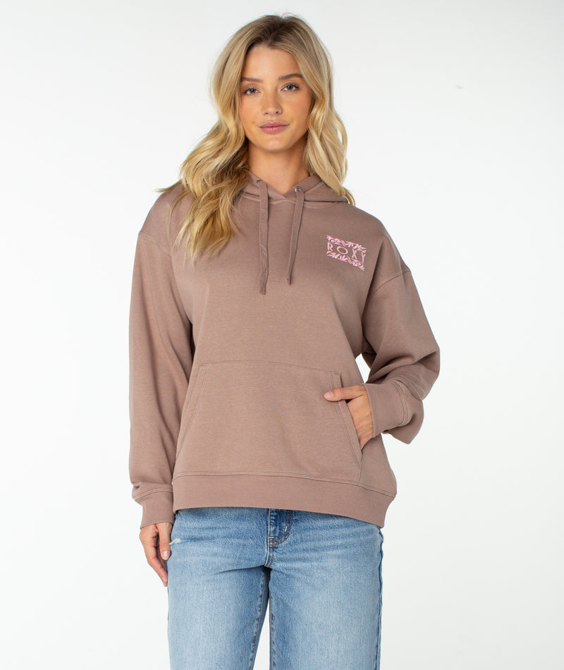 A woman with long blonde hair wears the Roxy Wailea Hoodie in Ginger Snap Palm Poster, featuring a pink graphic on the chest, paired with blue jeans as she stands against a plain white background.